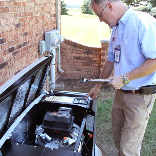 A technician in a light blue shirt and tan pants inspecting an open Generac standby generator while holding a clipboard.